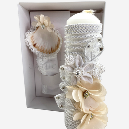 The image features a beautifully designed 3-piece baptism candle set, including a candle, a towel, and a shell. The candle is adorned with white fabric, a braided nylon rope, small white flowers, and rhinestone embellishments, giving it an elegant and sacred look. The towel and shell complement the set, creating a cohesive design perfect for a baptism ceremony.